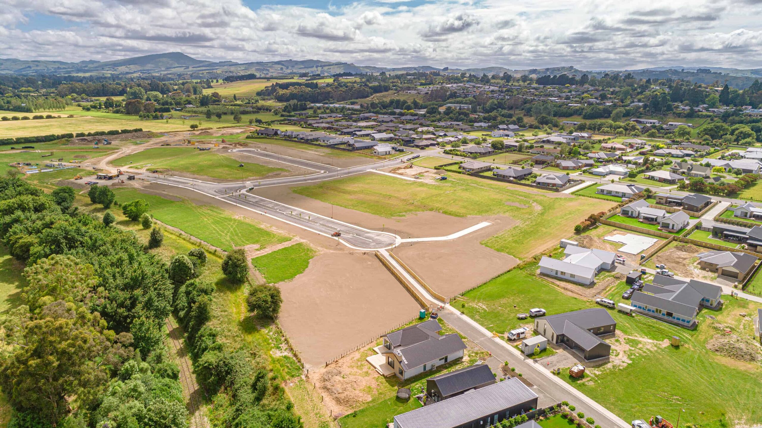 Aerial view of a suburban residential construction site with roads in place and scattered houses, surrounded by greenery and hills in the distance.