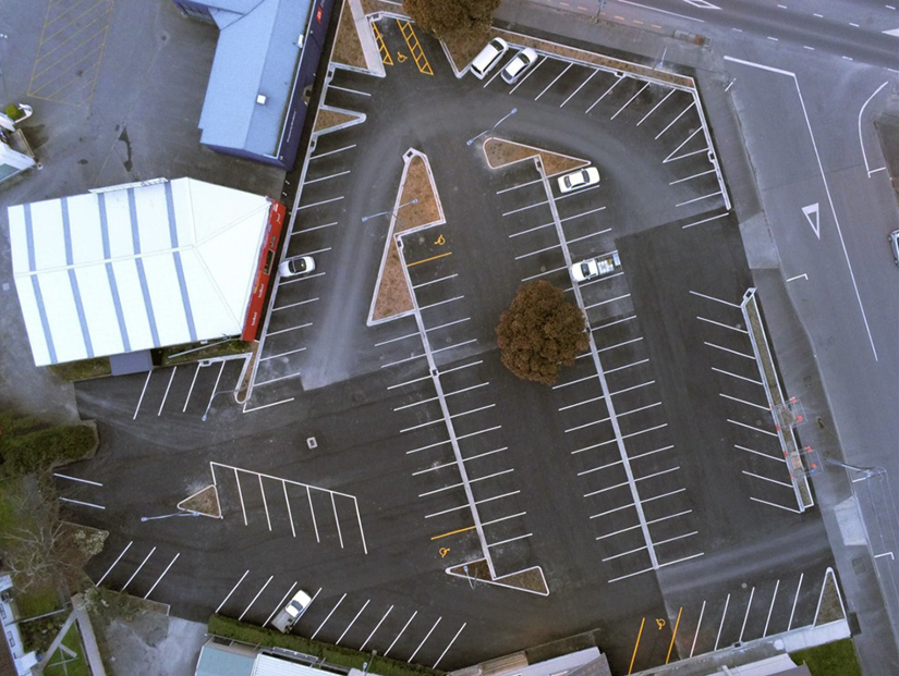 Aerial view of a nearly empty parking lot with a few cars, lined spaces, and some surrounding buildings and roads.
