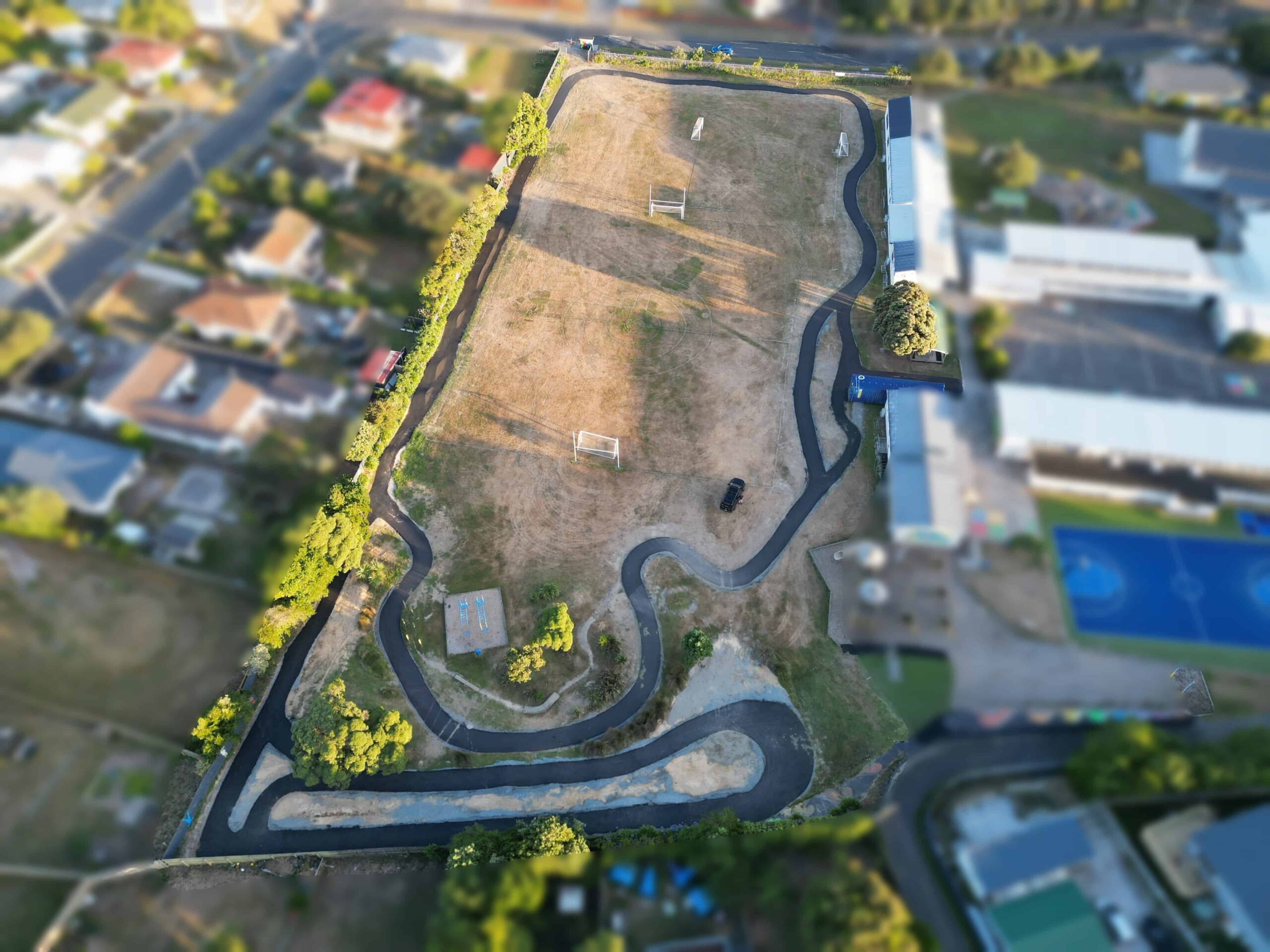 Aerial view of a fenced area featuring a winding paved path around a grass field with soccer goals, surrounded by residential houses and a sports facility.
