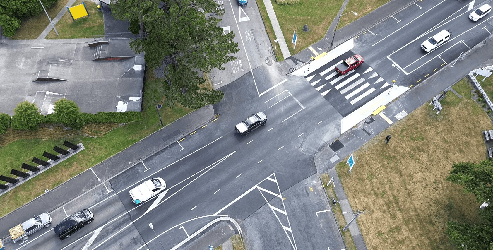 Aerial view of a multi-lane intersection with several cars, a pedestrian crossing, and surrounding greenery.