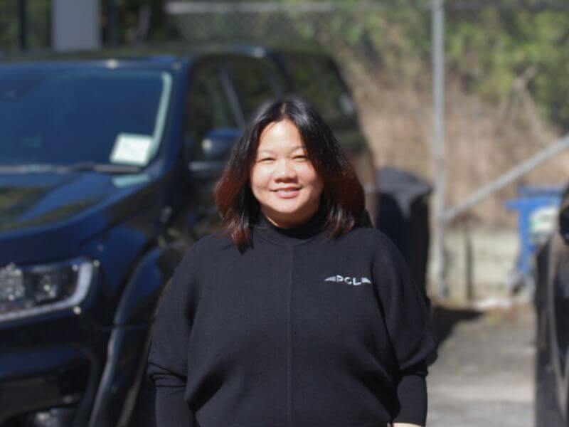 a person wearing a black sweater stands in front of a dark colored vehicle in an outdoor setting.