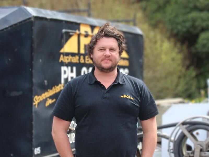 a man standing outdoors, wearing a black polo shirt with a logo, with construction equipment and greenery in the background.