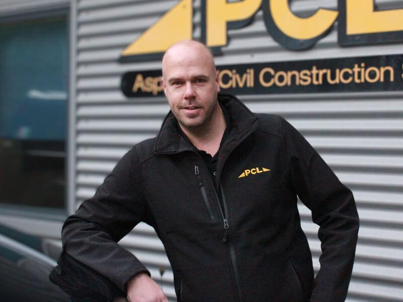 man standing in front of a building with a sign partially showing the name "pcl." he is wearing a black jacket with a yellow "pcl" logo. the background is corrugated metal siding.