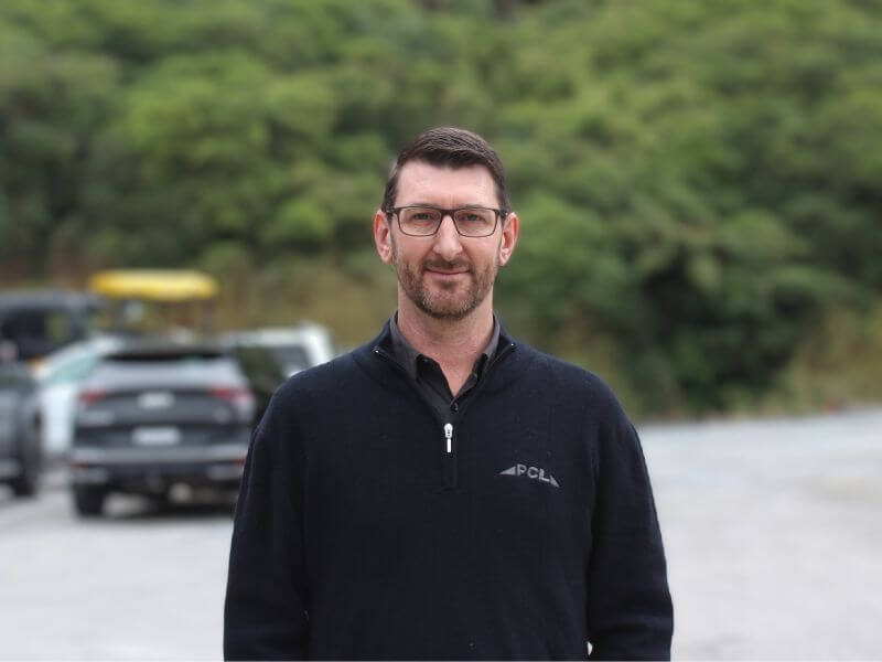 man with glasses and short hair wearing a dark sweater stands outdoors with trees and cars in the background.