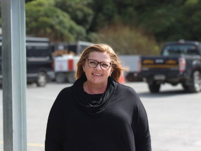a woman with glasses and wearing a black top is standing outdoors. there are trucks and greenery in the background.