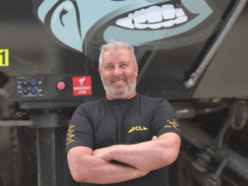 a man with short hair and a beard smiles with arms crossed, standing in front of equipment with an emergency stop button and a logo overhead.