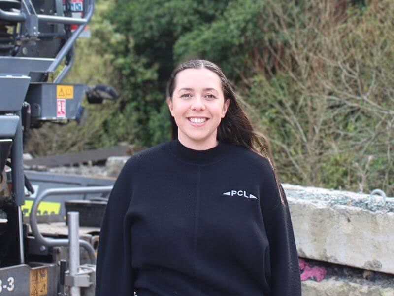 a person wearing a black sweater with the "pcl" logo stands outdoors near machinery and trees, smiling at the camera.
