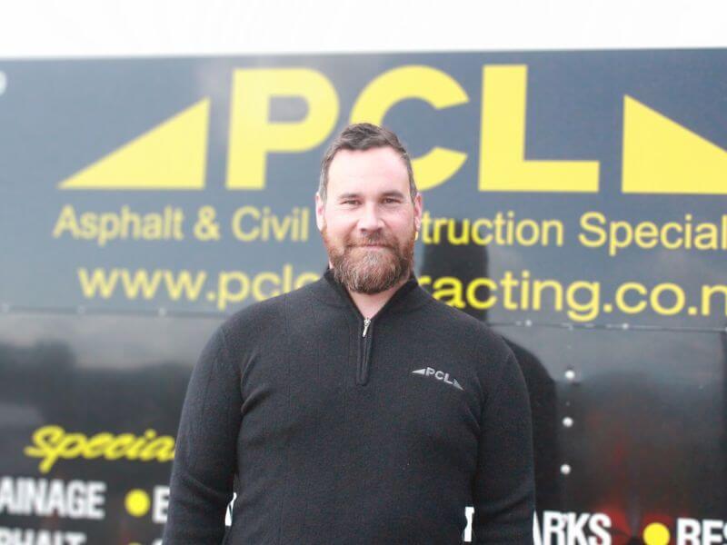 a man with a beard and black pcl sweatshirt stands in front of a pcl asphalt & civil construction specialists sign.