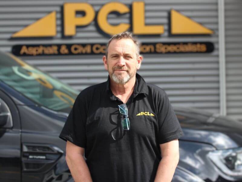 a man wearing a black shirt with a pcl logo stands in front of a vehicle and a sign that reads "pcl asphalt & civil construction specialists.