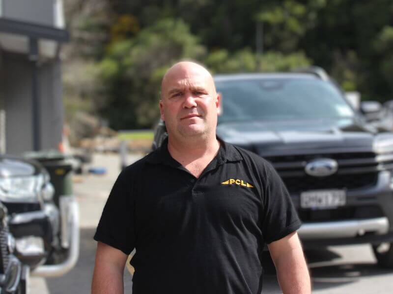 a man with a shaved head wearing a black pcl shirt stands in front of parked vehicles on a sunny day.