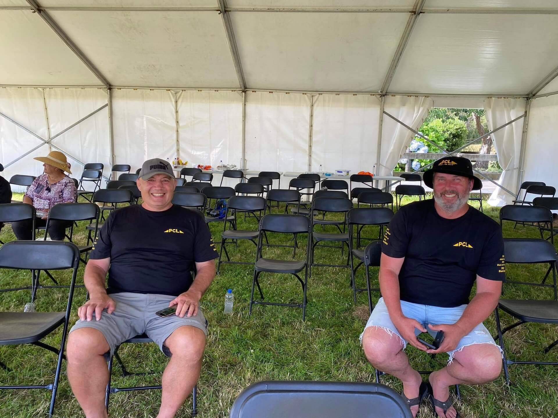 two men sitting in a tent with black folding chairs arranged in rows. each man is wearing a black shirt and a hat, smiling at the camera. a third person is visible in the background on the left.