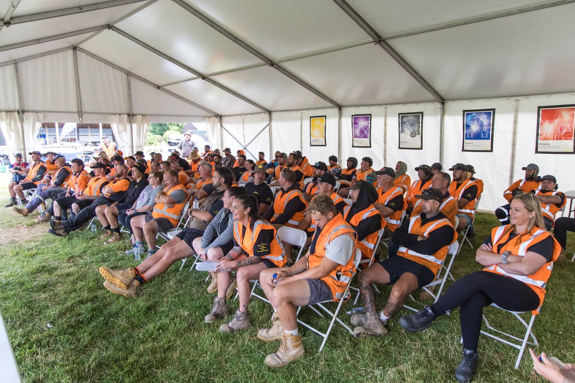 a large group of people wearing orange safety vests sit on white folding chairs under a large tent, attentively listening to a speaker outside the frame.