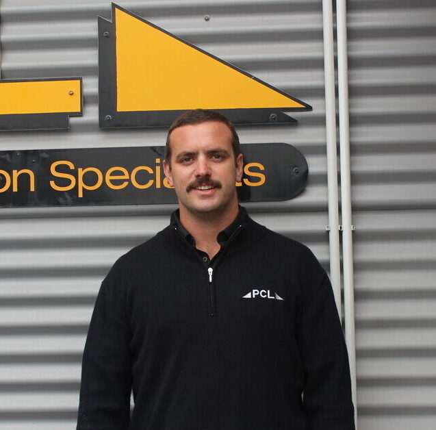 a man with a mustache stands in front of a corrugated metal wall, wearing a black pcl branded sweater.