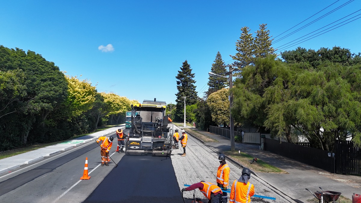 workers in orange vests operate machinery to pave a road under a clear blue sky, with trees lining both sides.
