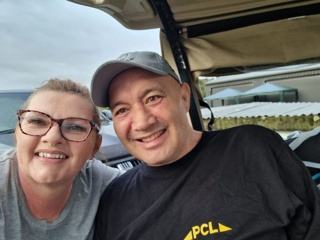two people smiling at the camera, seated in an open vehicle. the person on the left is wearing glasses, and the person on the right is wearing a cap and a black shirt.