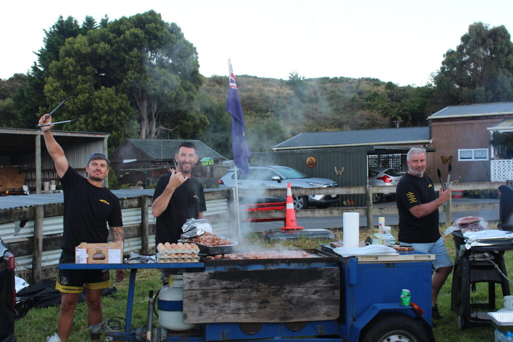 three men standing by a large outdoor grill cooking meat. two are holding utensils, and smoke rises from the grill. trees and buildings are in the background.