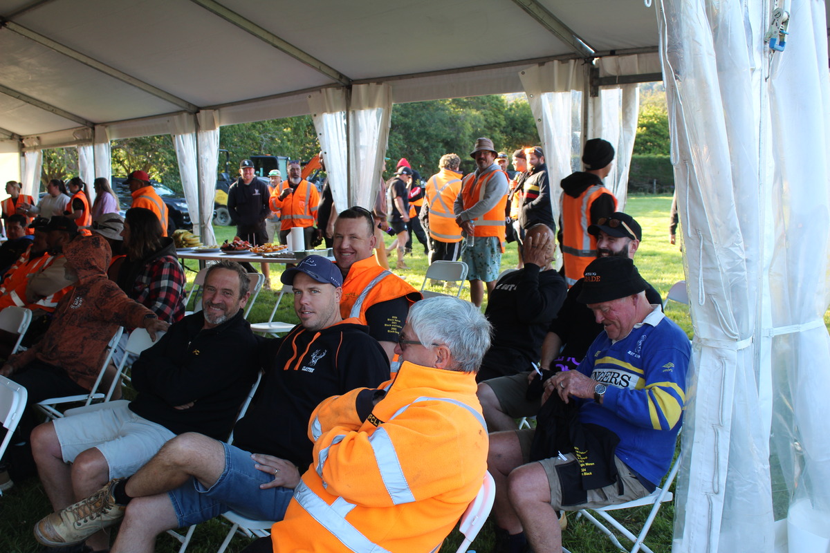 people in bright orange and casual clothing sit and chat under a white outdoor tent, with more people standing and mingling in the background.