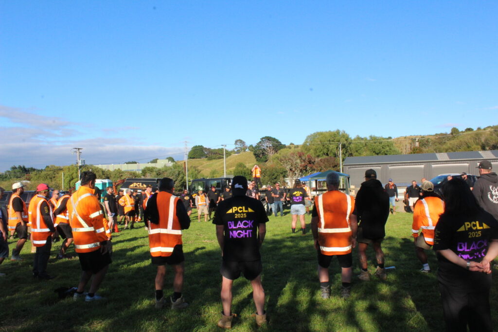 a group of people wearing orange safety vests and black shirts stand in a circle on grass under a clear blue sky.