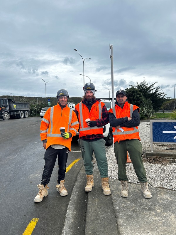 three construction workers in orange vests and hard hats stand on a roadside, each holding a beverage, with vehicles and cloudy skies in the background.