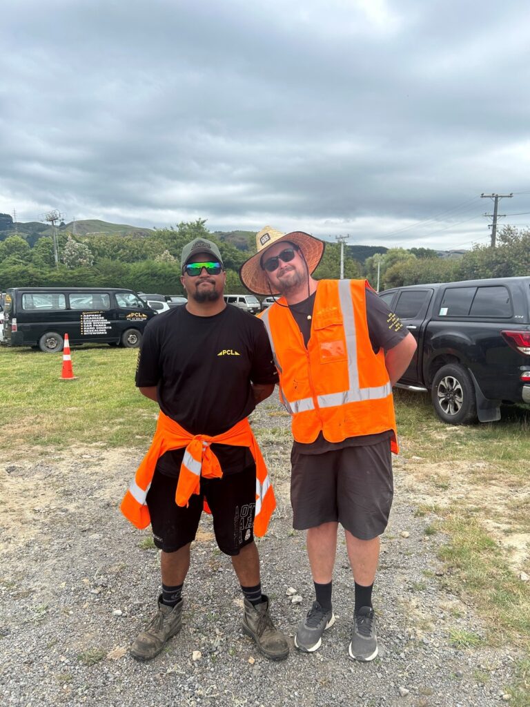 two men wearing orange safety vests, one with a hat, stand outdoors on a gravel area. vehicles and traffic cones are visible in the background. cloudy sky overhead.