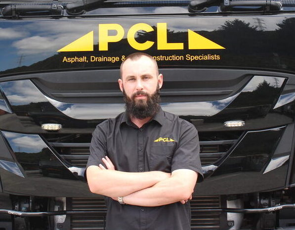 man with a beard, wearing a black shirt with pcl logo, stands with arms crossed in front of a large black vehicle branded with pcl signage.