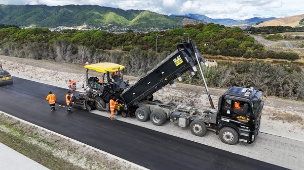 road construction workers operate machinery laying fresh asphalt, with a truck unloading pavement material onto the road, surrounded by hills and vegetation.