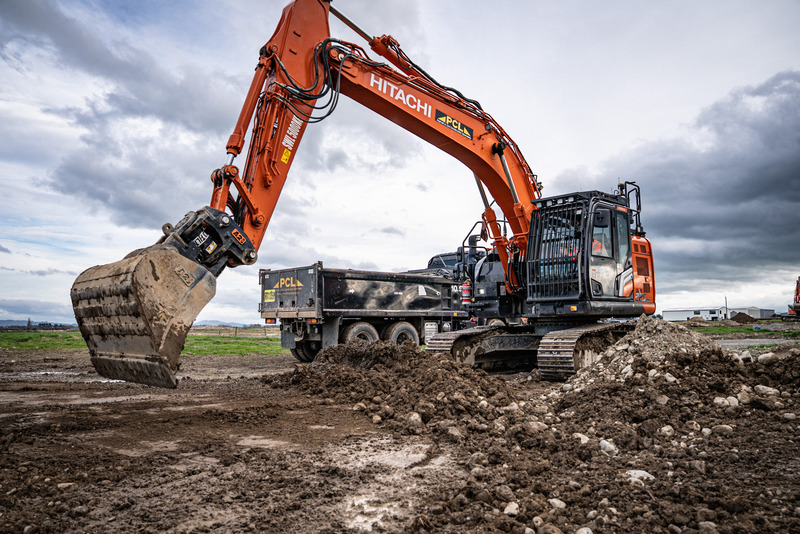 an orange hitachi excavator loads dirt into a black dump truck at a construction site under a cloudy sky.