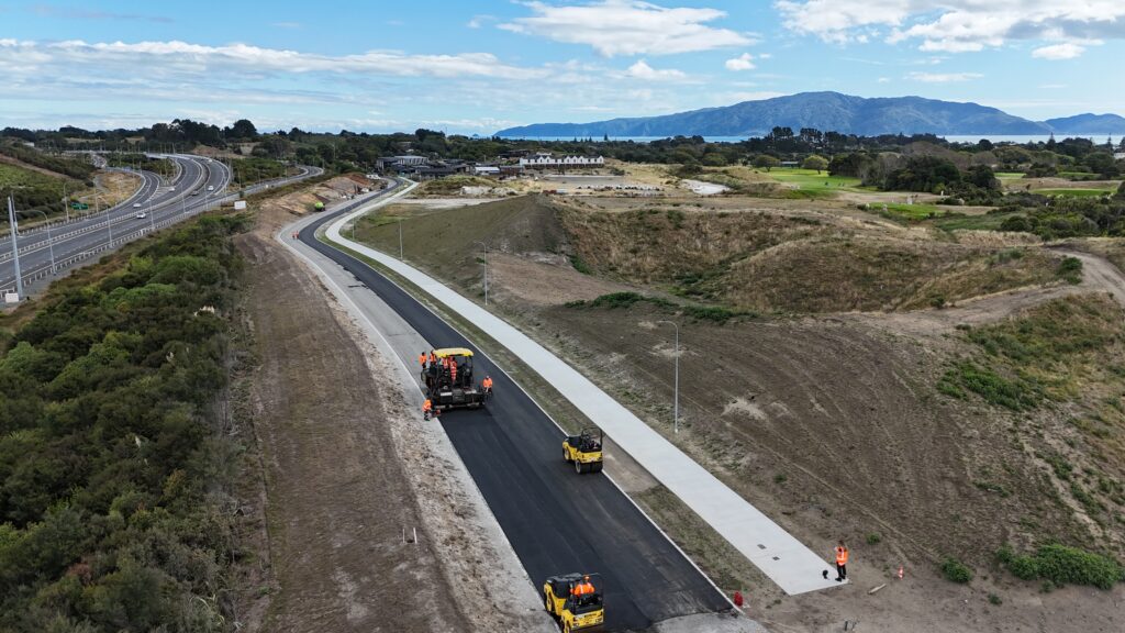 aerial view of construction workers paving a new asphalt road with machinery in a rural area, with hills, greenery, and distant mountains visible.