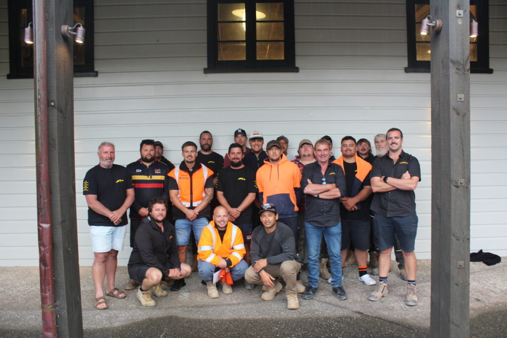 a group of men, some in hi vis workwear, pose for a photo outside a white building with two black framed windows.