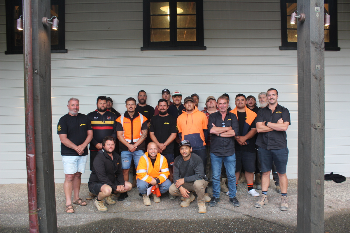 a group of men, some in hi vis workwear, pose for a photo outside a white building with two black framed windows.