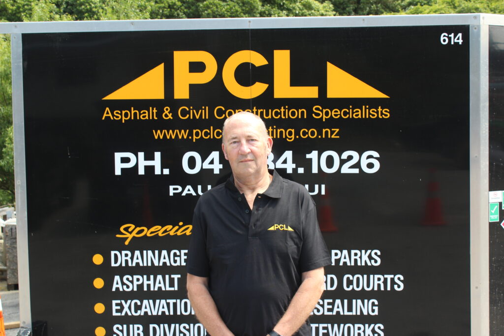 a man in a black pcl shirt stands in front of a black pcl asphalt & civil construction specialists sign with contact details and a list of services.