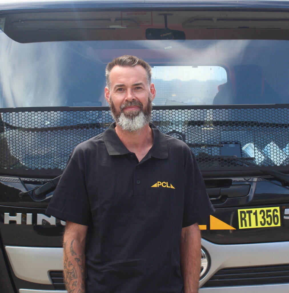 a man with a beard stands in front of a hino 500 truck, wearing a black polo shirt with "pcua" written on it.