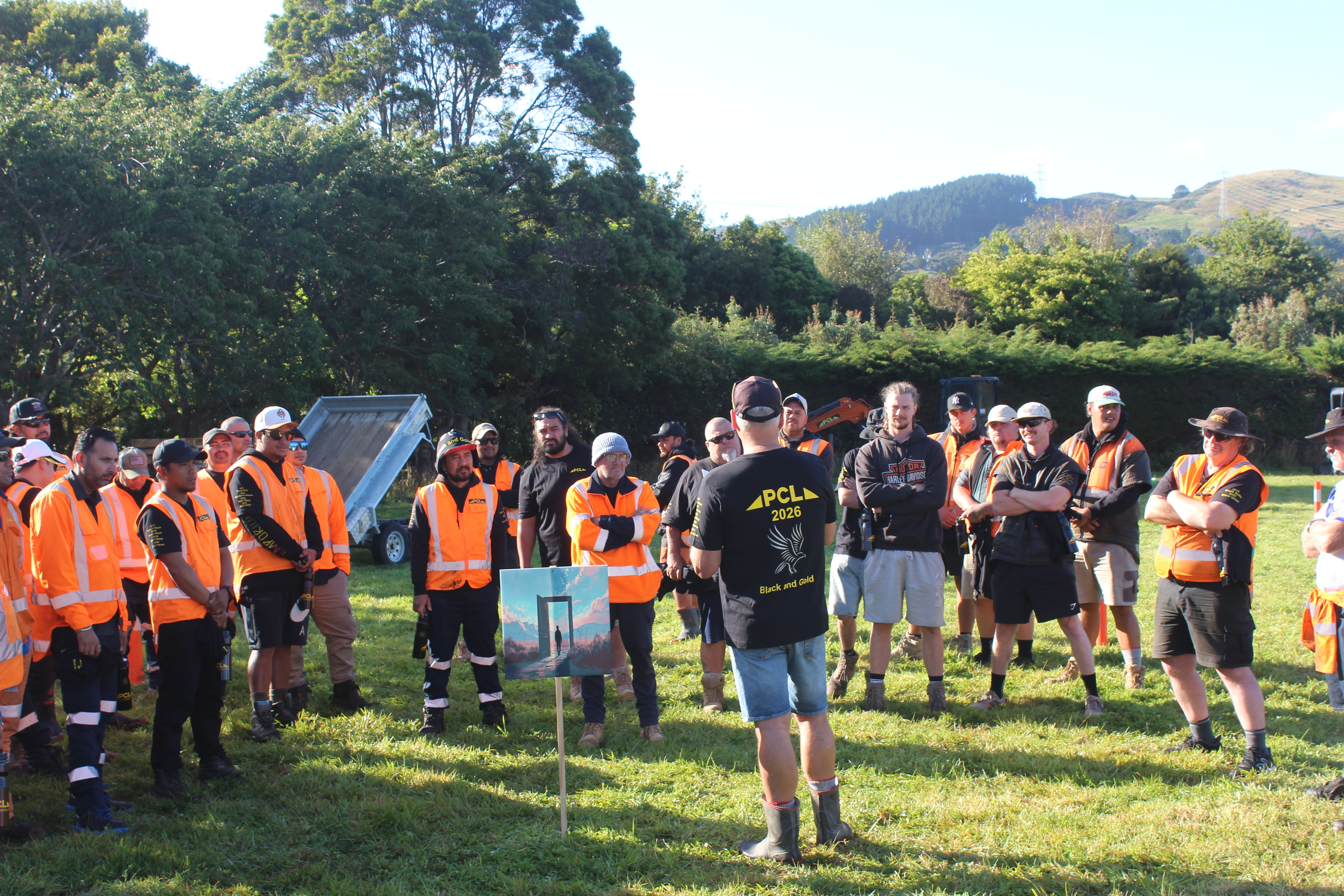 a group of people in orange safety vests listens to a person speaking outdoors; trees and hills are visible in the background.