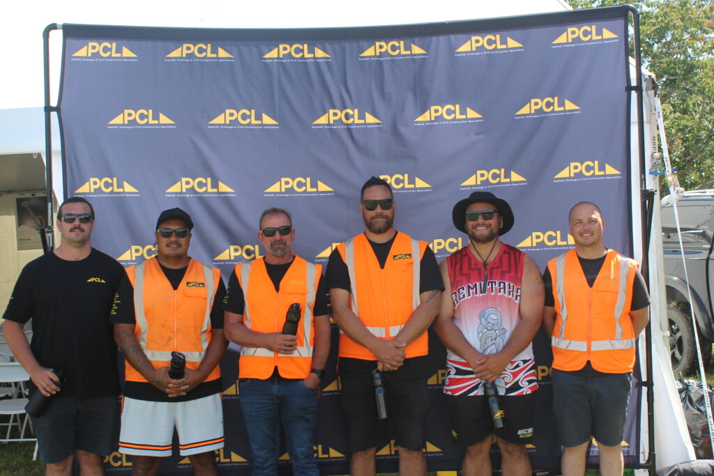 six men wearing orange safety vests stand in front of a pcl branded backdrop outdoors, posing for a group photo.