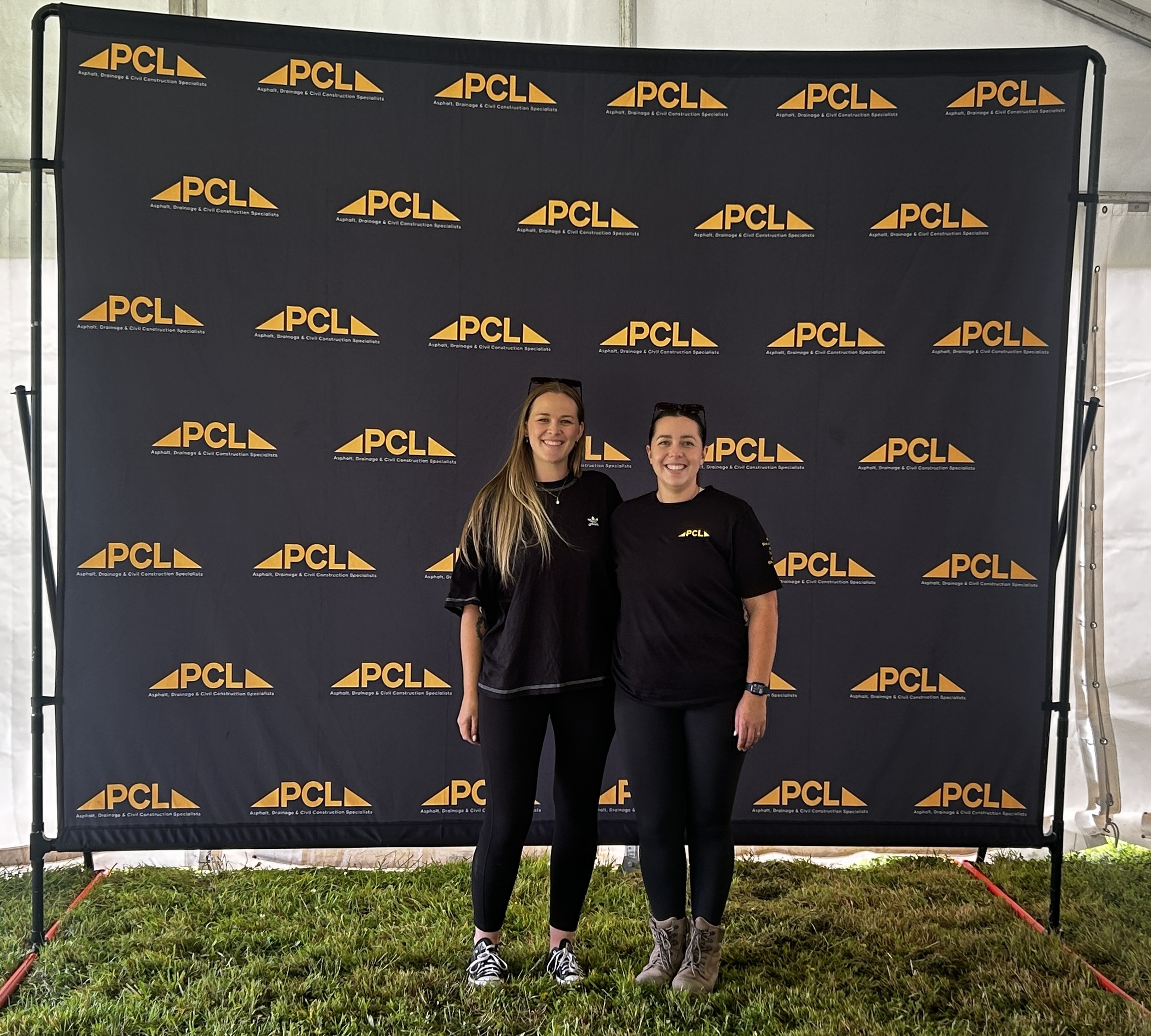 two people stand side by side, smiling in front of a black backdrop with repeated yellow "pcl" logos at an outdoor event.