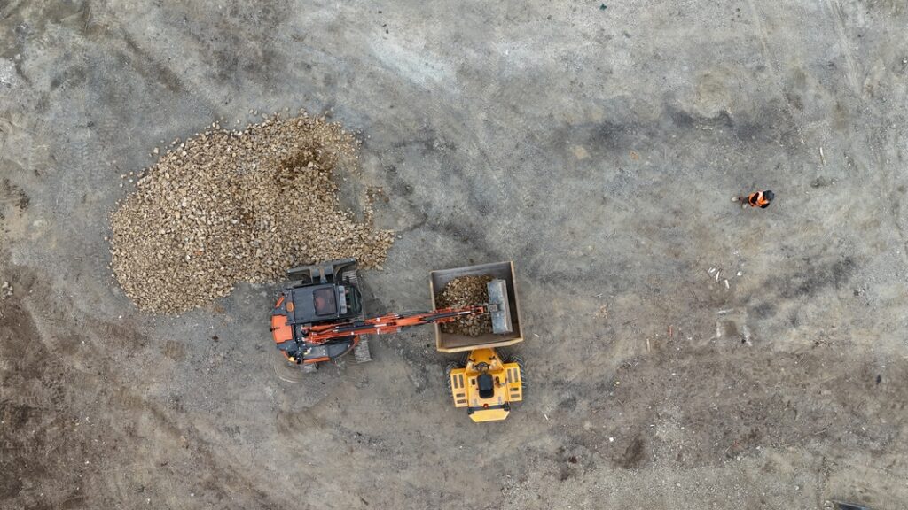 aerial view of two construction vehicles moving rocks on a dirt site, with a worker in an orange vest standing nearby.