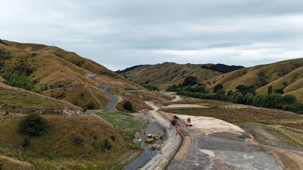 earthmoving equipment and construction workers are working on a rural road surrounded by rolling, grassy hills under an overcast sky.